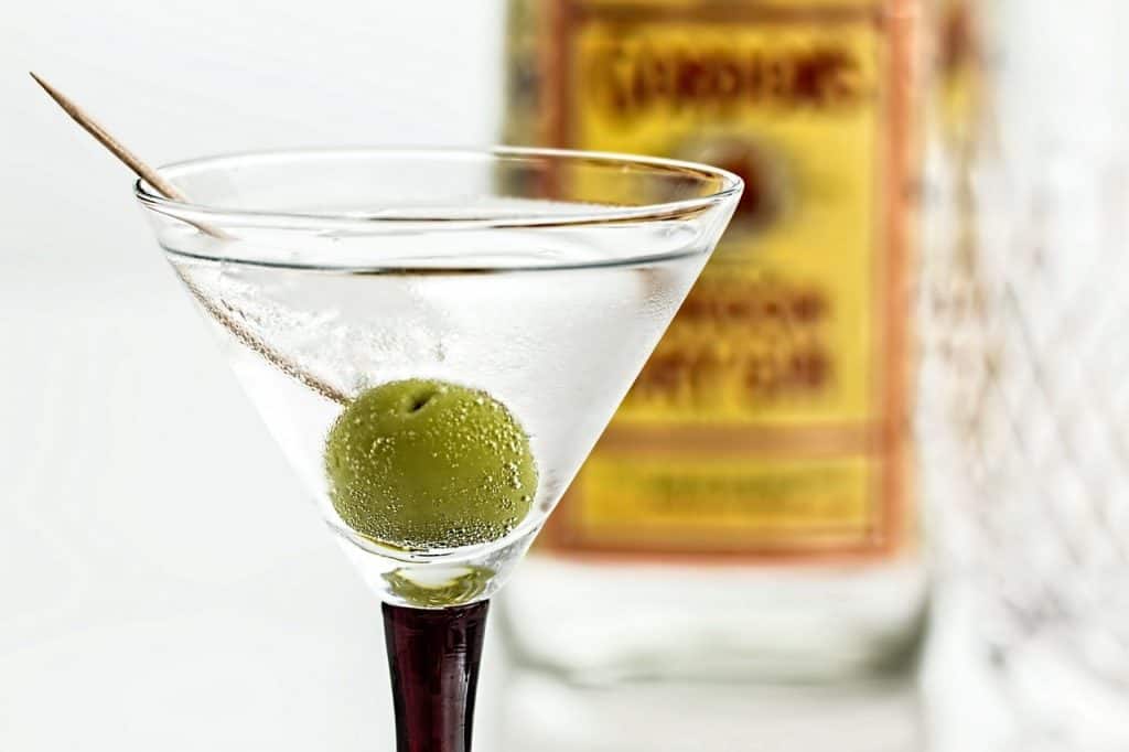 An up close view of a martini cocktail with an olive. The background shows a blurry bottle of Gordon's gin.