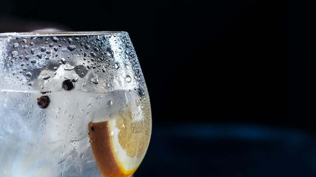 An upclose view of a bubble glass containing gin, a wedge of lemon, and peppercorns.