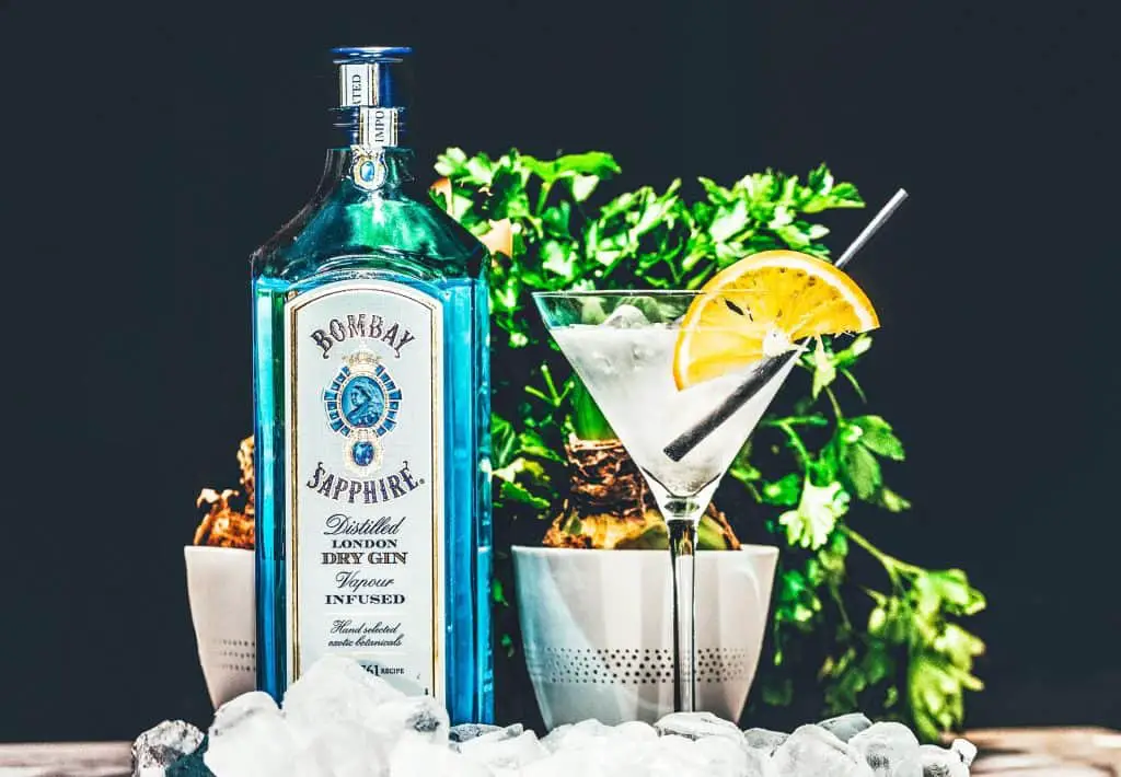 Up close bottle of Bombay Sapphire next to a cocktail glass that has a black straw and large slice of lemon in it.