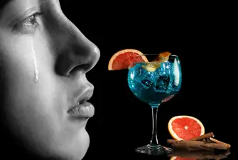 A person in black and white with a tear running down their face next to a blue gin cocktail.