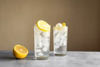 Two delicious Tom Collin cocktails on a marble worktop. The glasses are ice cold with a slice of lemon on the side.