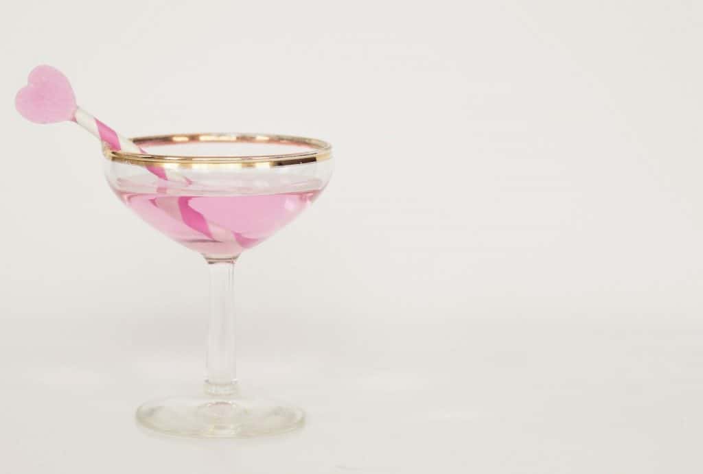 A stunning looking cocktail glass with a golden rim that has pink gin inside of it together with a white and pink stirring stick that has a pink love heart on top.