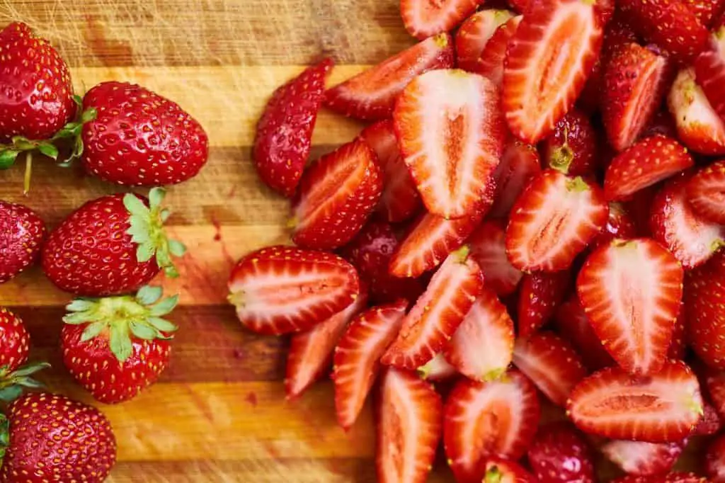 A large number of strawberries on a wooden cutting board. The strawberries on the left are whole while on the right side they are halved.