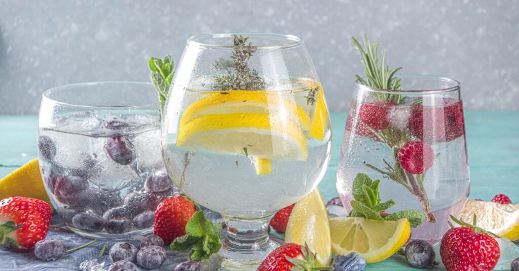 Three full glasses of gin, each filled with something different. The left glass has berries and ice, the middle glass lemon and fresh herbs, and the right glass contains berries and rosemary.